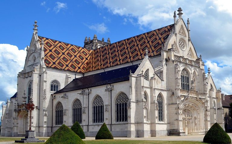 A large Gothic-style church with ornate white stone facades, pointed arches, and stained-glass windows stands as a true hymne de pierre. Its steep roof features colorful, geometric patterned tiles. Manicured bushes and a blue sky with clouds complete the scene.