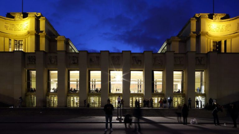 A night view of the Palais de Chaillot in Paris, illuminated with golden lights against a deep blue sky. People stand and walk on the steps in front, their silhouettes framed by the Théâtre’s large, glowing windows.