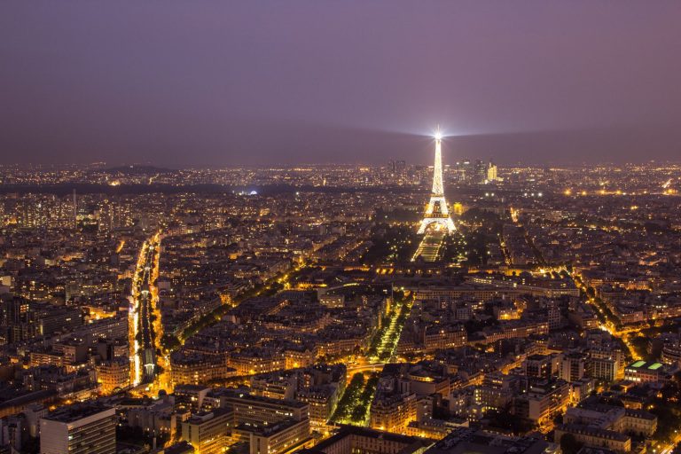 A nighttime aerial view of Paris shows the city illuminated by streetlights. The Eiffel Tower stands at the center, aglow and beaming a spotlight, while landmarks like Hôtel Renoir Paris blend into the vibrant avenues stretching toward the hazy horizon.