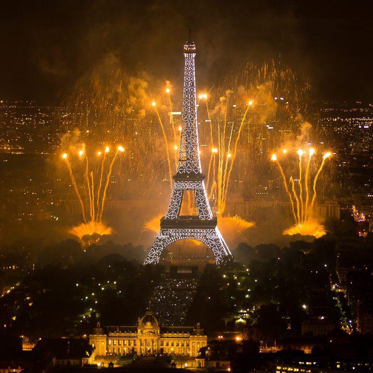 The Eiffel Tower in Paris is illuminated at night, with bright golden fireworks exploding behind it. Nearby, the Hôtel Renoir Paris sits among the glowing cityscape, as the warm light reveals streets and buildings surrounding the tower.