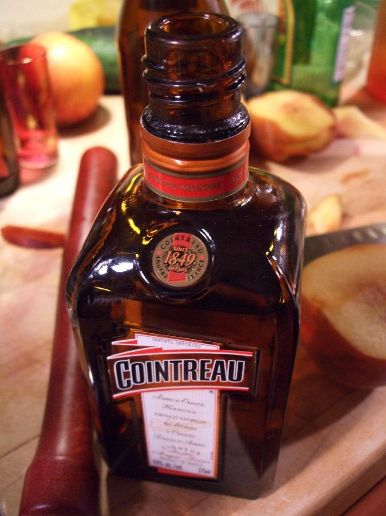 A close-up of a brown glass Cointreau liqueur bottle, the iconic marque mondiale, sits on a wooden counter. Its label and orange cap stand out amid blurred cocktail-making tools like fruits, a red muddler, and a knife.