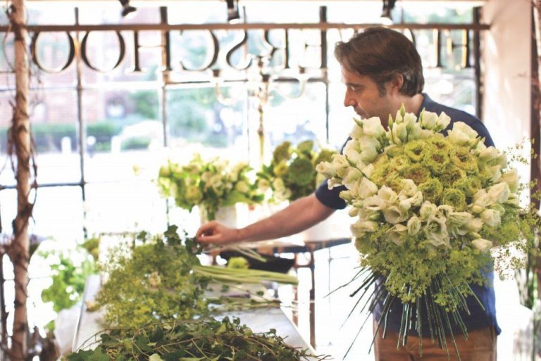 A man arranges fleurs in a bright, rustic shop inspired by Christian Tortu. He holds a large, round bouquet of white lilies and green foliage, with more flowers spread on a wooden table as sunlight streams through the windows.
