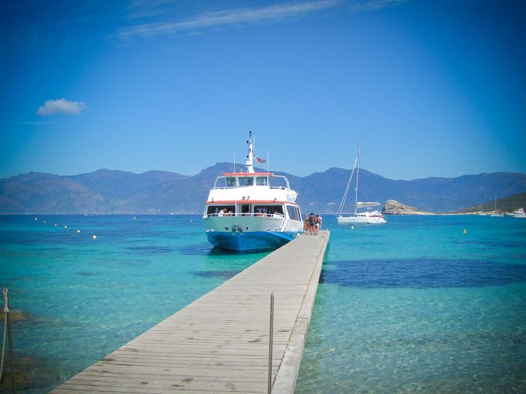 A white and blue ferry is docked at the end of a wooden pier over clear turquoise water, with people boarding. In the background are mountains under a bright blue sky and another boat anchored further out. The scene is calm and sunlit.