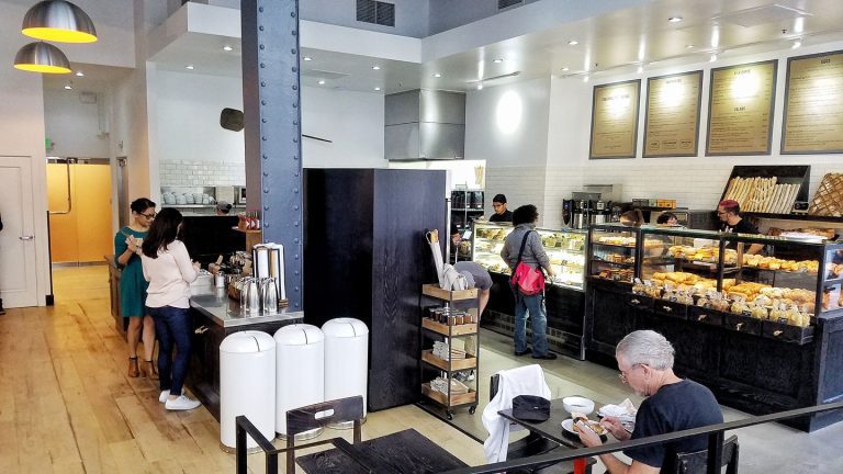 A modern bakery café inspired by Pascal Rigo, famed San Francisco boulanger, with customers ordering at a counter of pastries. Staff work behind glass cases, while a man sits upfront. The space features light wood floors, white walls, and industrial accents.