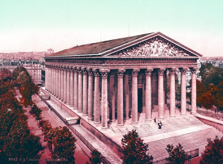 A neoclassical building with tall Corinthian columns, broad steps, and a sculpted pediment. Trees line the walkway as people stand near the entrance, while the Parisian cityscape—echoing the gloire of Napoléon 1er—spreads under a pale sky.