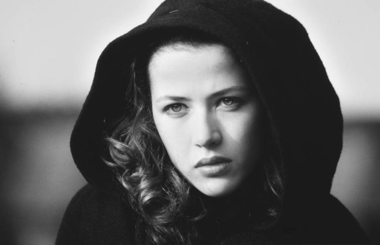 Black and white close-up of a young woman with wavy hair, her serious gaze and dark hooded coat echoing the poise of Sophie Marceau. The blurred background draws focus to her contemplative expression, inviting quiet bonheurs.