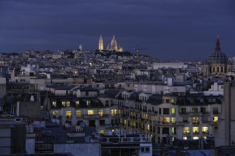 A twilight cityscape of Paris shows rows of buildings with glowing windows. The illuminated Sacré-Cœur Basilica sits atop Montmartre hill, while a glimpse of the Champs Elysees and a construction crane add to the vibrant scene under a cloudy sky.