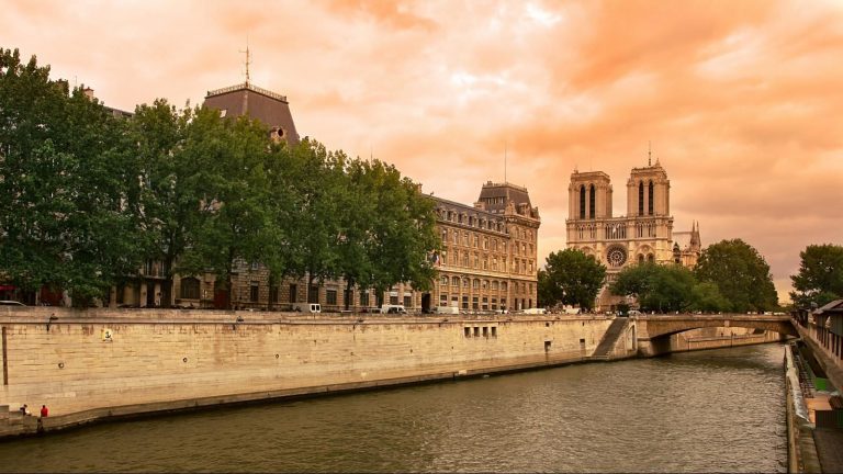 A view of the Seine River in Paris at sunset, with leafy trees and historic stone buildings near Hotel Le Clos Medicis lining the left bank. The Notre-Dame Cathedral stands prominently beneath a dramatic orange sky.
