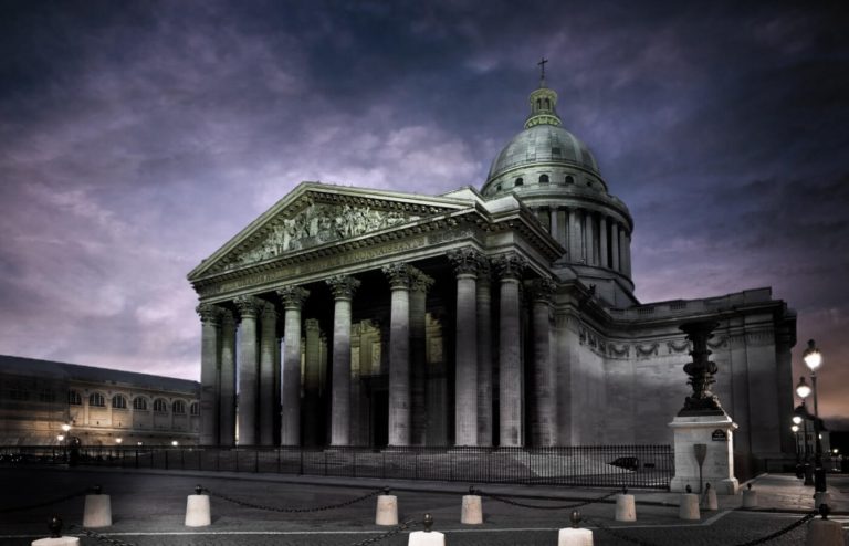 A grand neoclassical building with tall Corinthian columns and a large central dome stands under a cloudy evening sky near Hotel Moderne Saint-Germain, its spotlit facade overlooking cobblestoned streets in the heart of Paris.