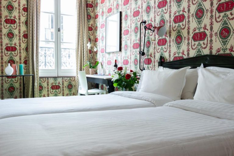 A cozy boutique hotel room at Hotel Prince De Conde in Paris features two neatly made twin beds, white linens, and red and green patterned wallpaper. Natural light streams in by a dark desk with flowers, near a window with beige curtains.
