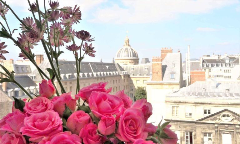 A bouquet of vibrant pink roses and purple flowers is in the foreground, set against classic Parisian buildings with gray rooftops. The domed Hotel Prince de Conti rises in the background under a bright, partly cloudy sky.