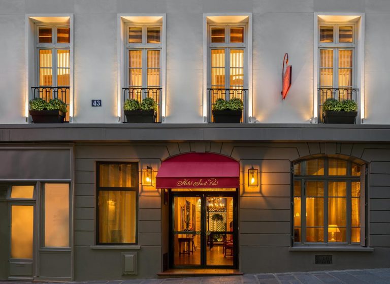 A warmly lit hotel facade at night features tall windows with black railings and flower boxes, a red awning over the entrance reading “Hotel Saint Paul” in white script, and a glowing wall sconce. The light beige building boasts elegant decorative trim.