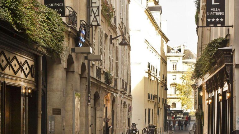 A narrow Parisian street lined with historic buildings; signs for boutique hotels like Résidence des Arts and Hotel Villa d'Estrees hang above, with greenery cascading from windows. Bicycles line the cobbled street, illuminated by sunlight.