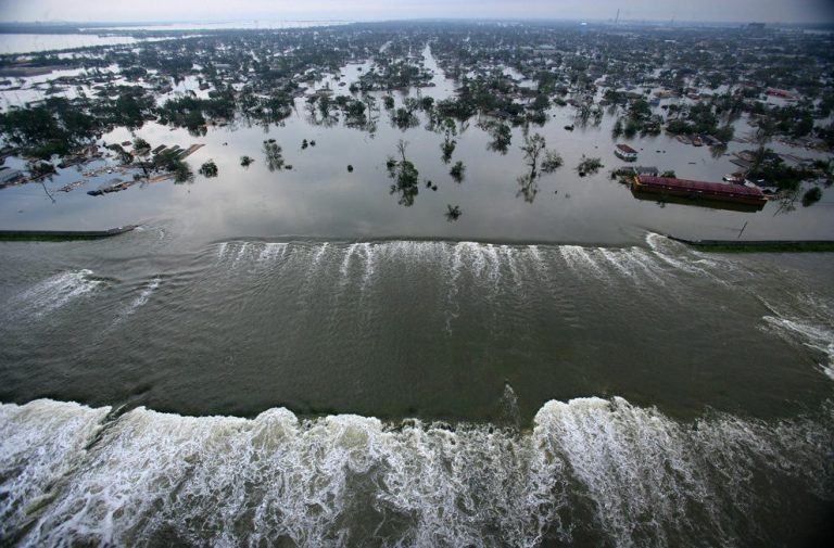 Aerial view of a flooded city with water pouring over a levee in the foreground. Neighborhoods, trees, and buildings—some known for their restauration architecturale—are submerged, with waters extending into the distance under a cloudy sky.
