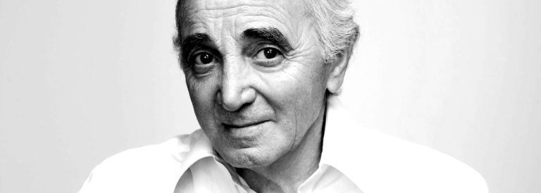 A black-and-white portrait against a soft, plain background of Charles Aznavour,as an older man with white hair and thick eyebrows, in a white collared shirt. He looks directly at the camera with a gentle, slight smile and laughter lines begining to crease his cheeks.