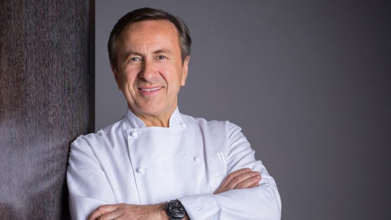 A smiling middle-aged chef with dark hair stands with arms crossed, wearing a white chef’s coat and a black watch. He stands against a dark wooden wall with a plain grey background, capturing a moment in the vie d'un chef.