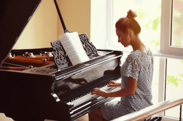 A woman in a light blue lace dress sits at a grand piano, playing music. Sheet music is open on the stand as she continues to apprendre le piano, bathed in the warm, soft glow of sunlight streaming through large windows.