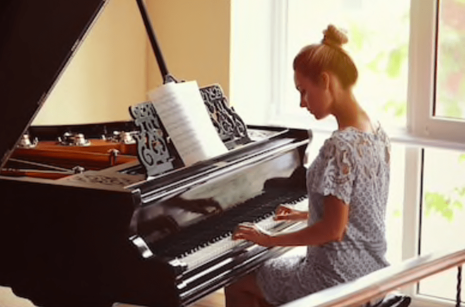 Une femme vêtue d'une robe en dentelle bleu clair est assise à un piano à queue et joue de la musique. Les partitions sont ouvertes sur le pupitre tandis qu'elle continue d'apprendre le piano, baignée par la lumière chaude et douce du soleil qui traverse les grandes fenêtres. 