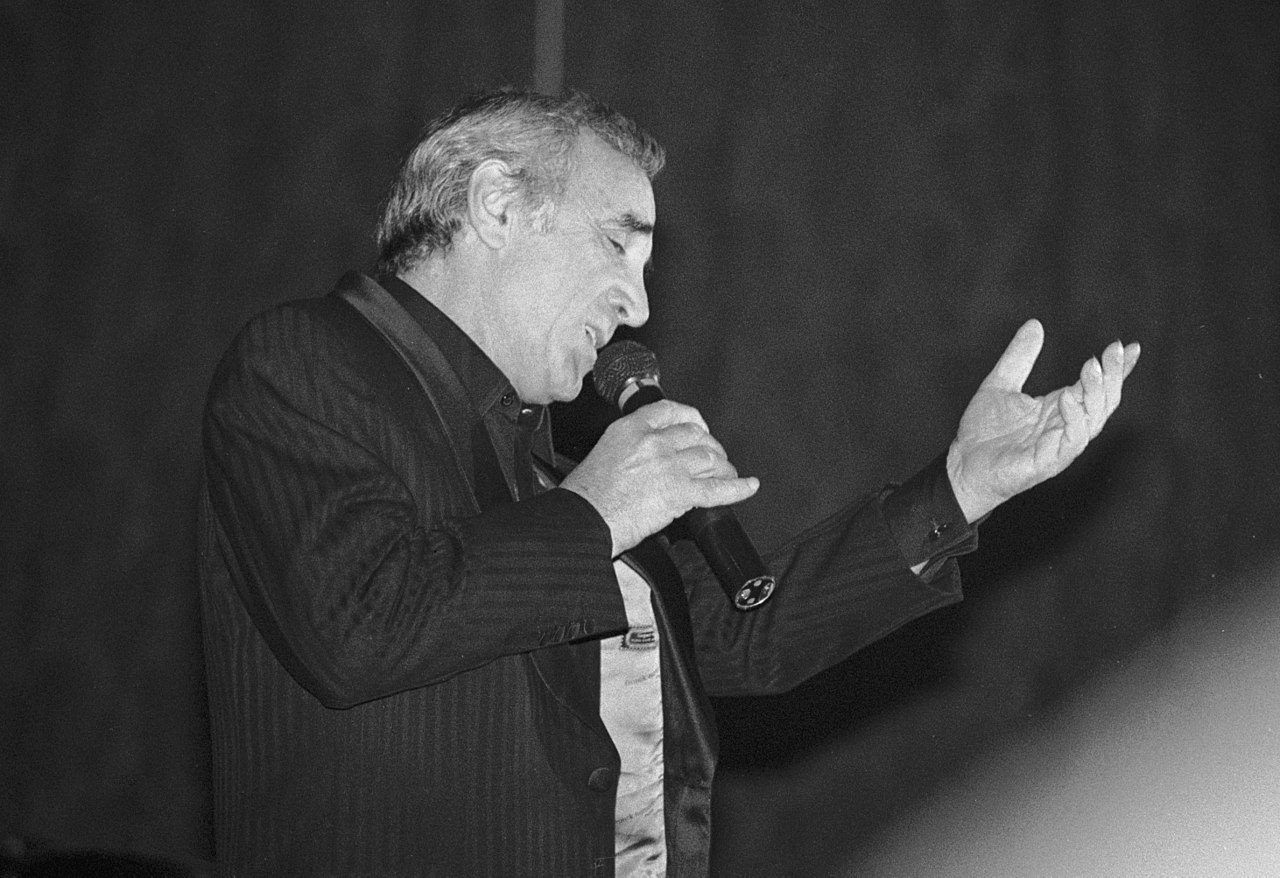 Photo en noir et blanc d'un homme âgé aux cheveux gris, vêtu d'un costume à rayures sombres, chantant avec passion, la main gauche levée, les yeux fermés. Véritable référence, il incarne l'esprit d'Aznavour à chaque note. L'arrière-plan est flou.  