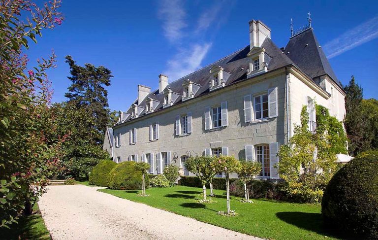 The grand Château de Détilly boasts pale stone walls and tall, white-framed windows, nestled beside a gravel driveway. Manicured bushes and small trees adorn the lush green lawn beneath a clear sky with wispy clouds.