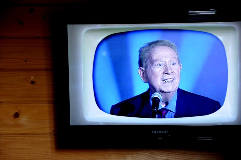 A television mounted on a wooden wall displays an older man with gray hair, evoking the âge d'or of chanson française, speaking into a microphone. He wears a dark suit, red tie, and blue shirt against a blue background on a retro-styled TV screen.