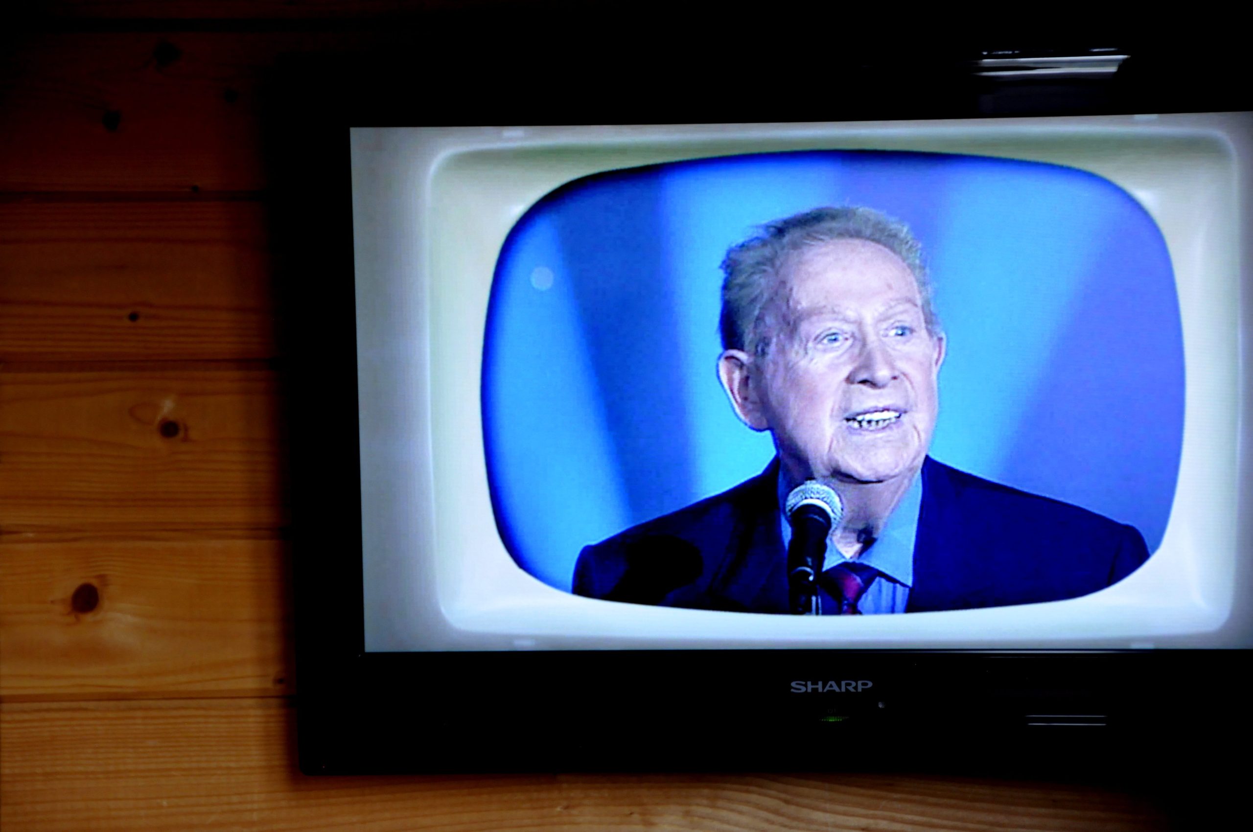 Un téléviseur monté sur un mur en bois montre un homme âgé aux cheveux gris, évoquant l'âge d'or de la chanson française, qui parle dans un micro. Il porte un costume sombre, une cravate rouge et une chemise bleue sur un fond bleu, sur un écran de télévision de style rétro. 