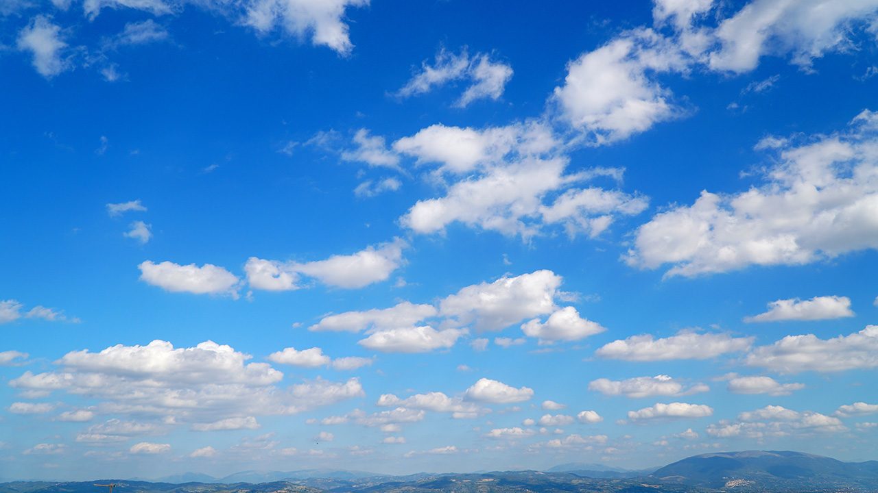 Un ciel d'un bleu éclatant parsemé de nuages blancs épars et cotonneux au-dessus de collines et de montagnes ondulantes lointaines. Le paysage brumeux en contrebas laisse entrevoir de nouveaux enjeux, créant un sentiment de profondeur et d'anticipation sous le ciel expansif. Aucune présence humaine ou animale n'est visible.  
