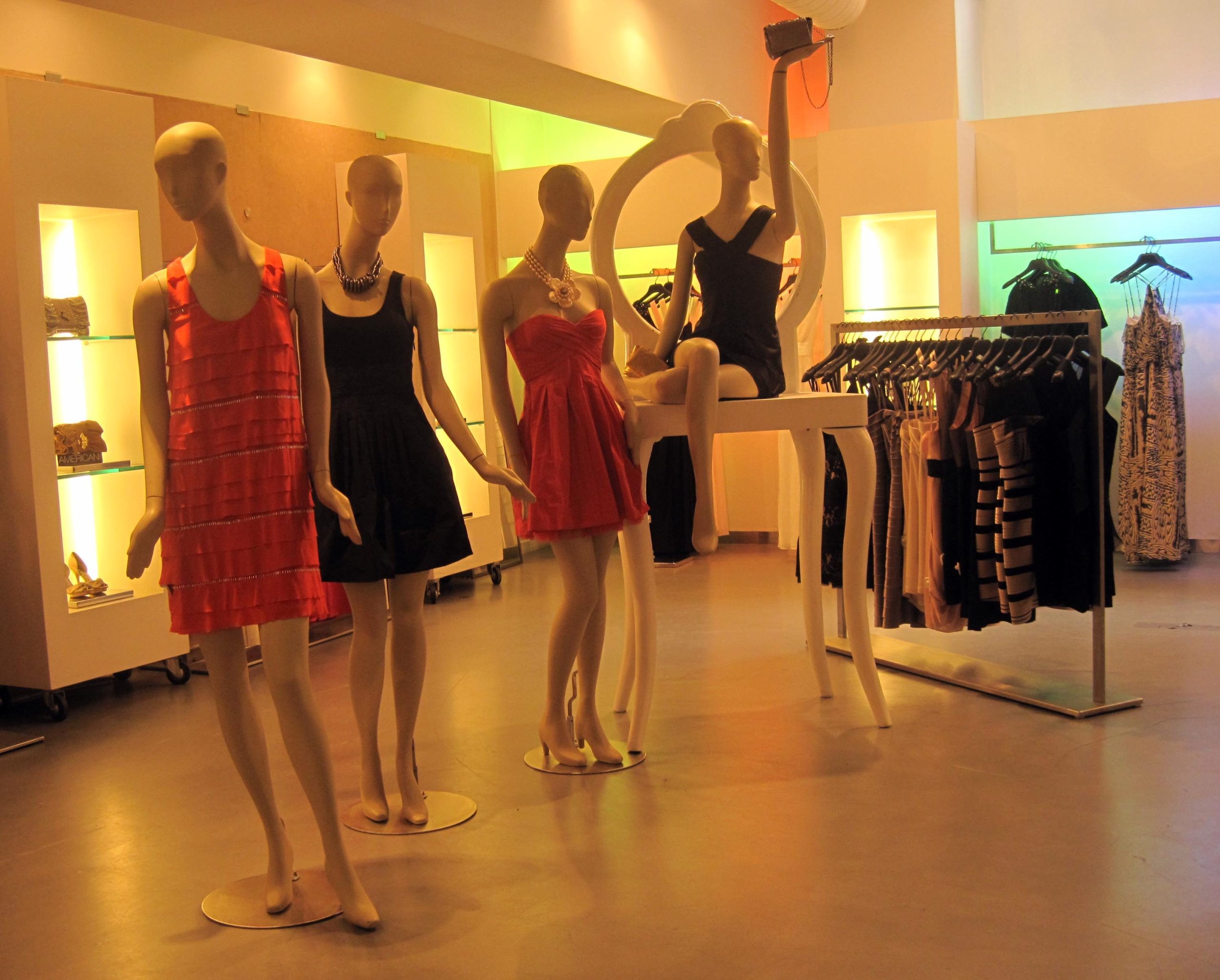 Quatre mannequins présentent des robes BCBG à la mode dans une boutique élégante et bien éclairée. Des portants de vêtements et des étagères illuminées bordent l'espace, créant une atmosphère moderne et élégante inspirée de Max Azria, chaleureuse et invitante. 