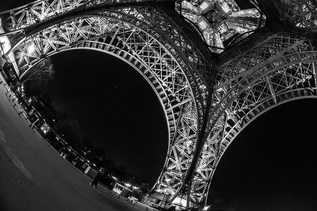 A black-and-white, low-angle view of the Eiffel Tower at night, its illuminated iron lattice arches curving dramatically through a fisheye lens—like musique française echoing across Paris, capturing the city’s timeless allure for the marché américain.