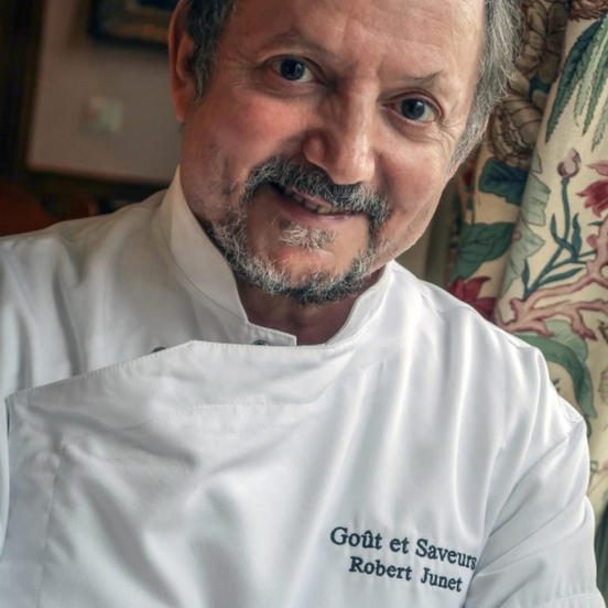 A smiling chef français with short, dark hair and a beard wears a white chef’s coat that reads Goût et Saveurs Robert Junet. He is indoors, standing near a window with floral curtains, the softly blurred background hinting at his départ à la retraite.