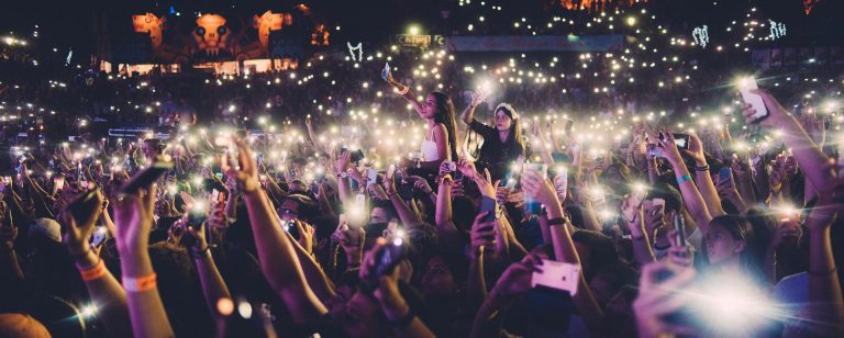 Au Festival musical du Languedoc-Roussillon, une foule nombreuse brandit des téléphones lumineux, créant ainsi une mer de lumières. Les gens applaudissent, sourient et prennent des photos au son d'une musique entraînante. Les lumières brillantes de la scène et les décorations colorées remplissent l'atmosphère énergique.  