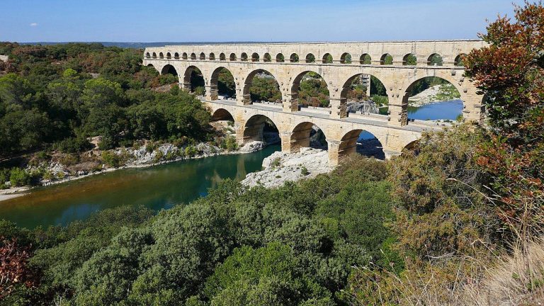Ancient Roman stone aqueduct bridge with multiple arches spans a river, surrounded by green trees and bushes under a clear blue sky. The bridges reflection is visible in the calm water below.
