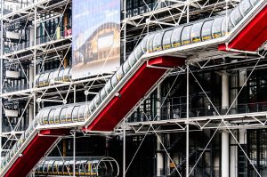 Exterior of the Centre Pompidou in Paris, showing exposed structural elements, red escalators in transparent tubes zigzagging across the façade, and an architectural banner. White steel beams and pipes create a geometric, industrial look.