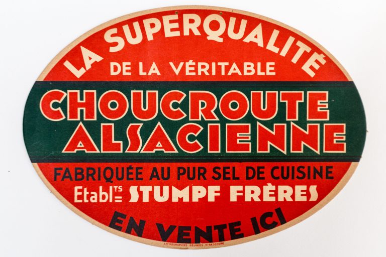 An oval vintage sign in red, green, and beige reads La Superqualité de la véritable Choucroute Alsacienne, celebrating authentic Sauerkraut. Bold, decorative text dominates the design and highlights its artisanal quality.