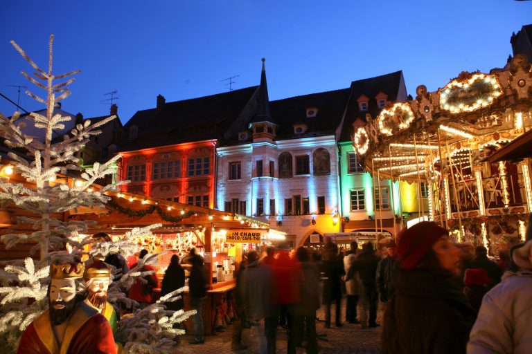 A vibrant Christmas market at dusk features festive lights, colorful illuminated buildings, a white Christmas tree, wooden stalls, a carousel, and a nativity scene. Crowds browse the stalls, capturing the magic of Christmas in France during the festive season.