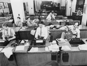 Black-and-white photo of a busy vintage newsroom. Men in dress shirts and ties work at desks with typewriters, telephones, and papers. Some are typing, one reads a newspaper, others are on the telephone. The room is filled with wooden furniture and office supplies.