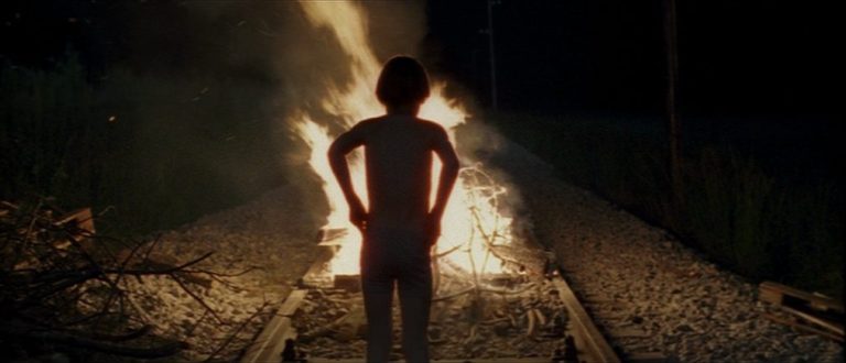 A person stands on a railroad track at night, silhouetted against a large, bright bonfire burning on the tracks ahead—a scene reminiscent of "Time of the Wolf," with flames illuminating scattered branches and stones in the darkness.