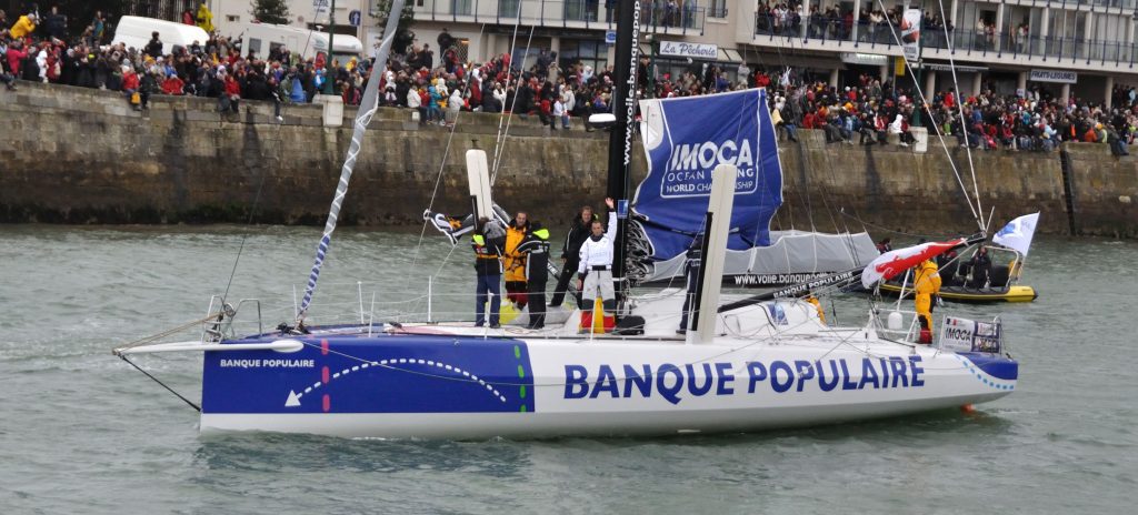 A blue and white sailboat labelled Banque Populaire sails past a crowd on shore. Three people stand on deck waving, with two holding raised paddles. The boat has a large blue sail and sponsor logos, and a small escort boat follows behind.