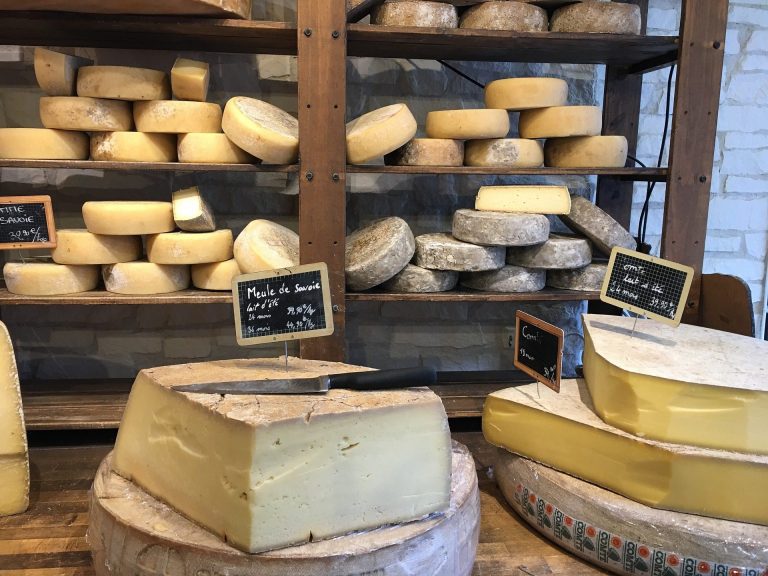 Sur les étagères sont empilées des meules et des rondelles de fromage français, certaines étiquetées par famille de fromage. Sur le comptoir en bois se trouvent de grands quartiers coupés, chacun marqué de petits signes noirs. Un couteau à fromage est posé sur l'un des quartiers de cette fromagerie accueillante.  