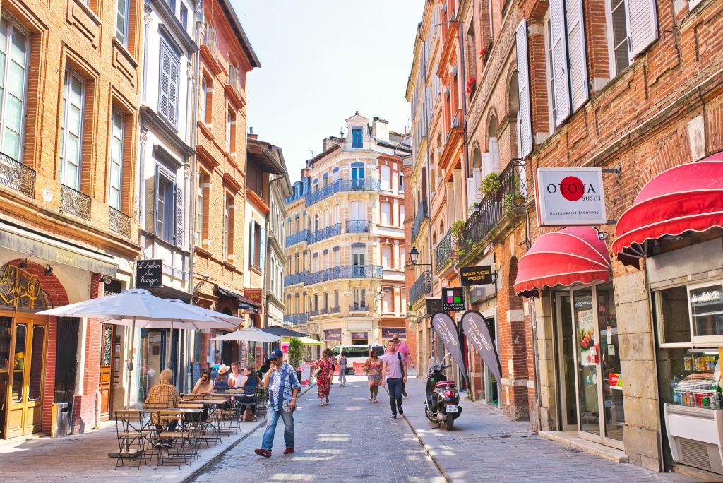 A lively European street scene shows people walking and dining at outdoor cafés. Tall, colourful historic buildings with red brick, awnings, and balconies line the cobbled street. Bicycles and a scooter are parked, and shop signs are visible. It is sunny and bright.