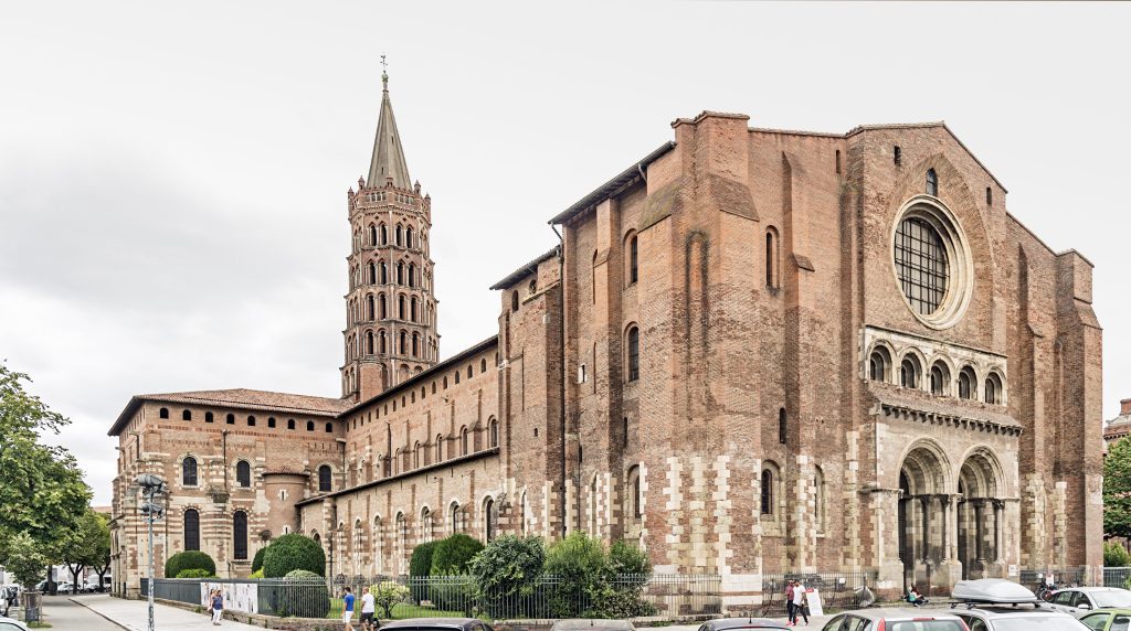 A large Romanesque brick church with arched windows, a tall octagonal bell tower, and a rose window above the main entrance. The façade features decorative columns, stone accents, and people walking in front. Trees line the left side of the scene.