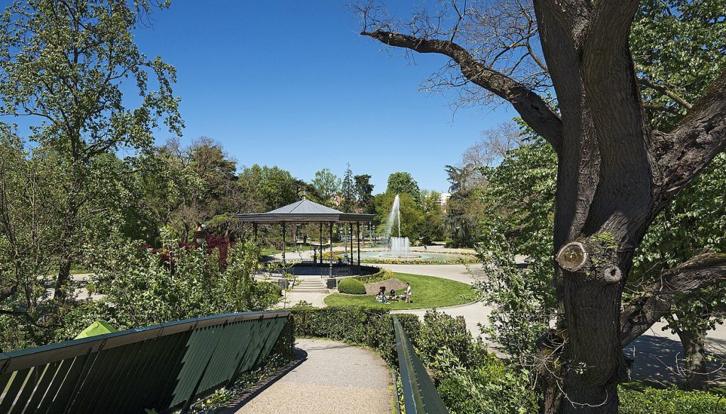 A lush park scene with a wooden railing leading down to a circular path. Trees and greenery surround a black gazebo and a tall fountain in the distance. People sit on benches, enjoying the sunny, clear blue sky.