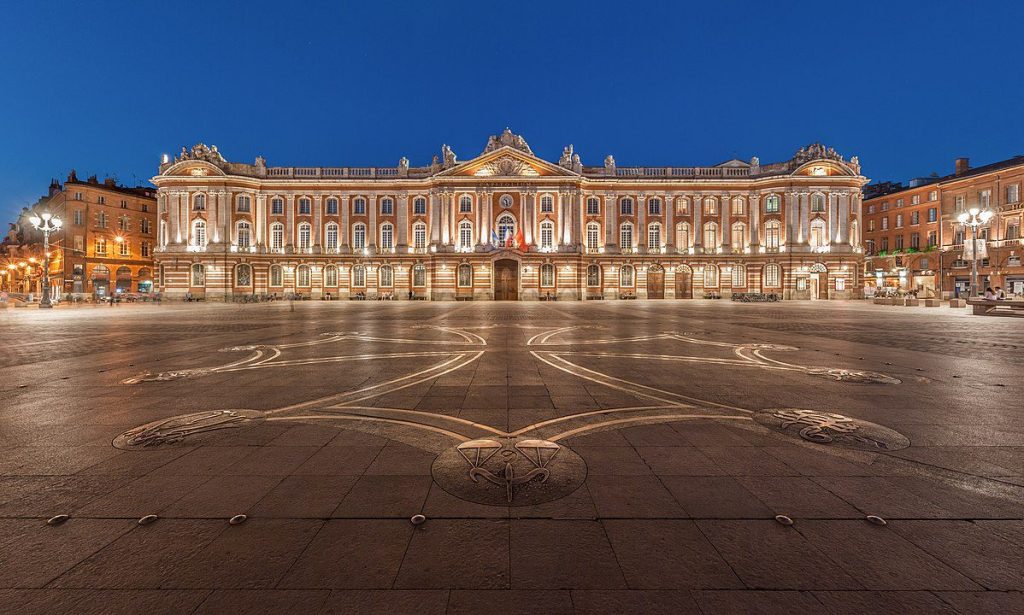 A wide, empty plaza at dusk with ornate stone patterns on the ground. In the background, a grand, symmetrical building with arched windows and decorative façades is brightly lit. Street lamps line the square, and a deep blue sky completes the scene.
