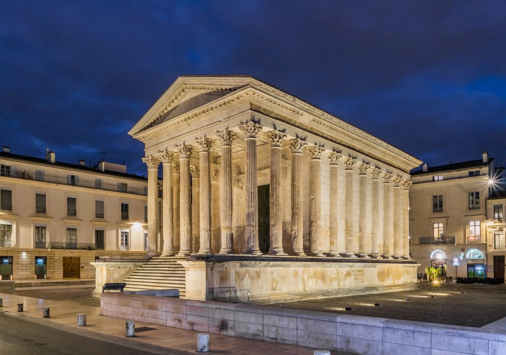 A well-preserved ancient Roman temple with tall Corinthian columns, a triangular pediment, and large stone steps, illuminated against a deep blue evening sky. The building stands in a city square surrounded by modern low-rise buildings.