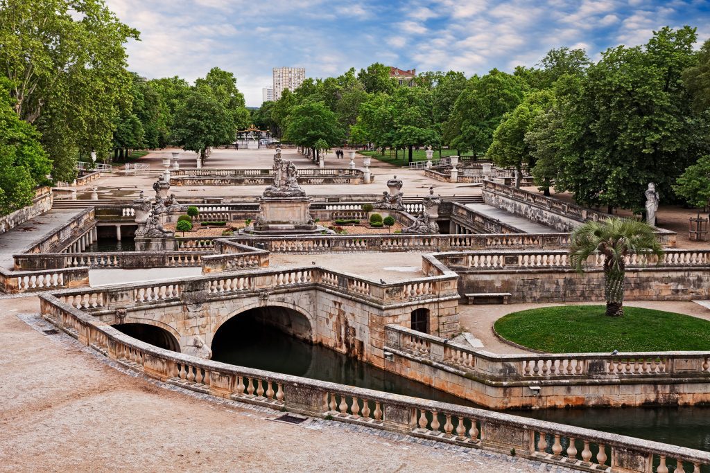 A formal stone garden with ornate balustrades, arched bridges over a canal, circular lawns, statues, and lush green trees, set under a partly cloudy sky. People walk in the distance; tall buildings peek through trees.