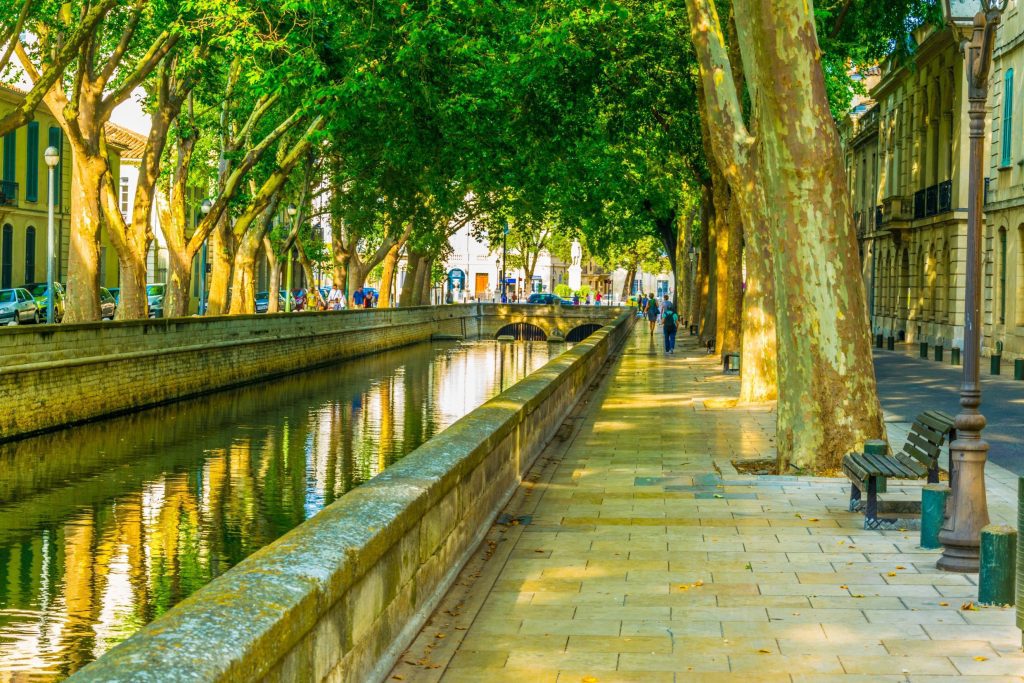 A sunlit, tree-lined canal runs beside a paved walkway with benches, old-style street lamps, and historic buildings. The lush green trees arch over the canal, creating dappled shade. A stone bridge crosses the water in the background, and people walk along the path.