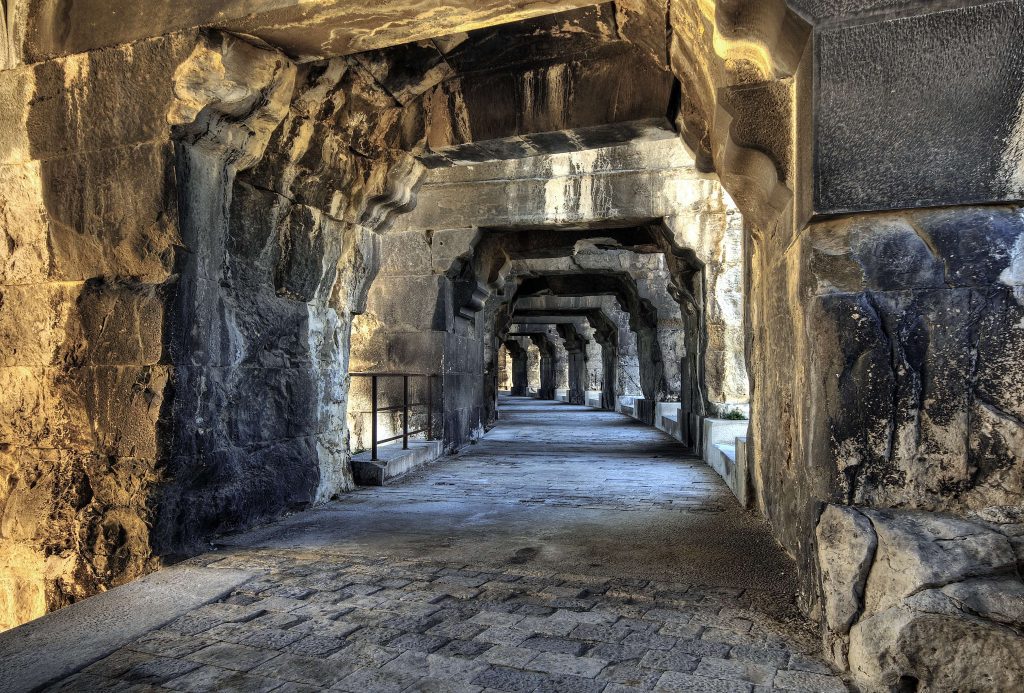 A stone corridor with repeating archways and rough-hewn walls, lit by sunlight from one side. The floor consists of uneven cobbles, and the passage appears ancient, stretching into the distance under angular stone arches.