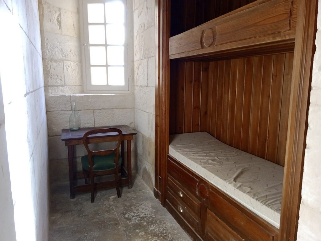A small, sparse room with stone walls features a wooden bunk bed with no bedding, a simple desk with a bottle and glass, and a wooden chair near a bright window letting in natural light. The floor is stone, and the room has a minimalist, old-fashioned feel.