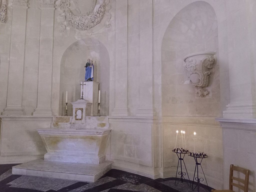 A small stone altar with a blue-robed statue of Mary holding baby Jesus stands in a light-coloured, ornate chapel. Five lit candles and votive holders sit on a black stand to the right. The walls feature carved decorative motifs and niches.