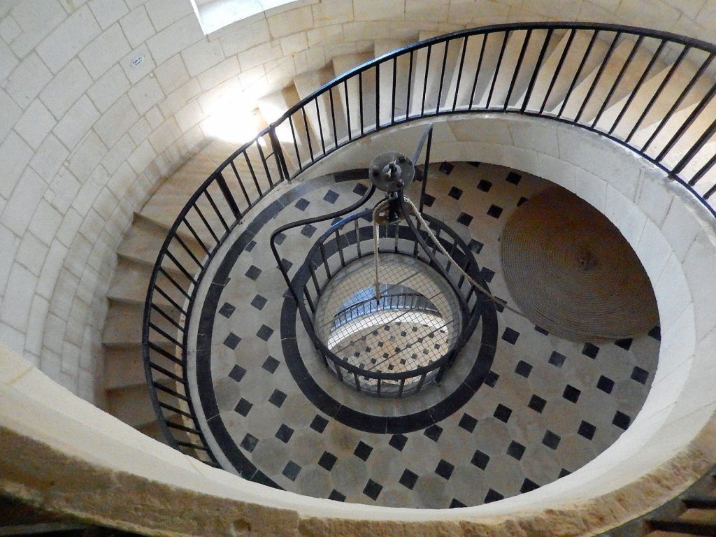 Spiral stone stairway curves around a central, black metal balustrade structure with a hanging lamp. The tiled floor below features a black and white diamond pattern, and circular rugs accent the space. The stone walls have a smooth, light-coloured finish.